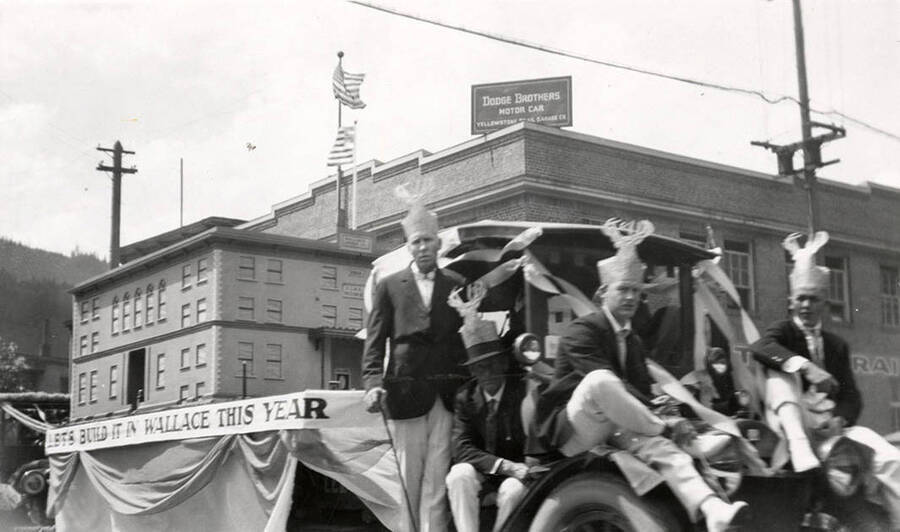 A group of men wearing hats driving a float in the Elks parade in Wallace, Idaho. The float has a sign on the side of it that says, "Let's build it in Wallace this year."