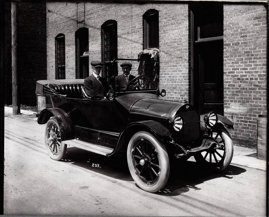Image shows two men sitting in a car, next to a building in Wallace. Caption on front: "Stone, Ed - Car Ed Stone and unidentified man in auto."