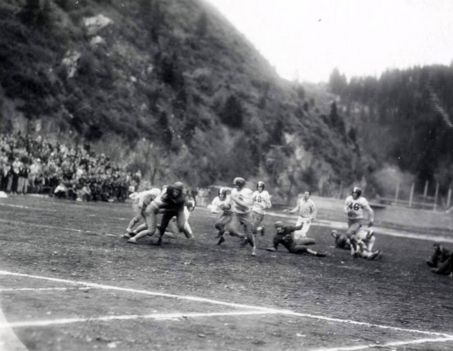 Football players playing in the Wallace-Sandpoint football game. A crowd of people sits on the sidelines, watching.