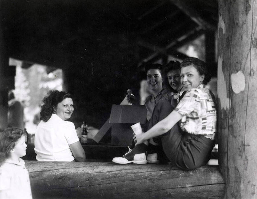 A group of people sitting on a log and at a picnic table during Wallace Jaycee's picnic in Pottsville, Idaho.