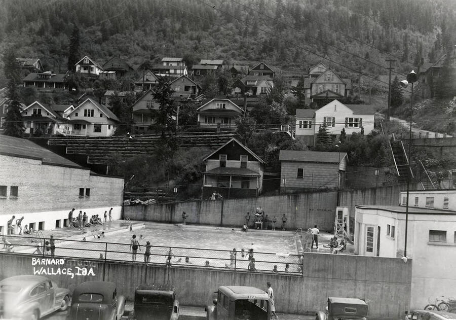View of the swimming pool in Wallace, Idaho. Many people are standing around the side of the pool and a few are swimming. Houses can be seen surrounding the swimming pool.