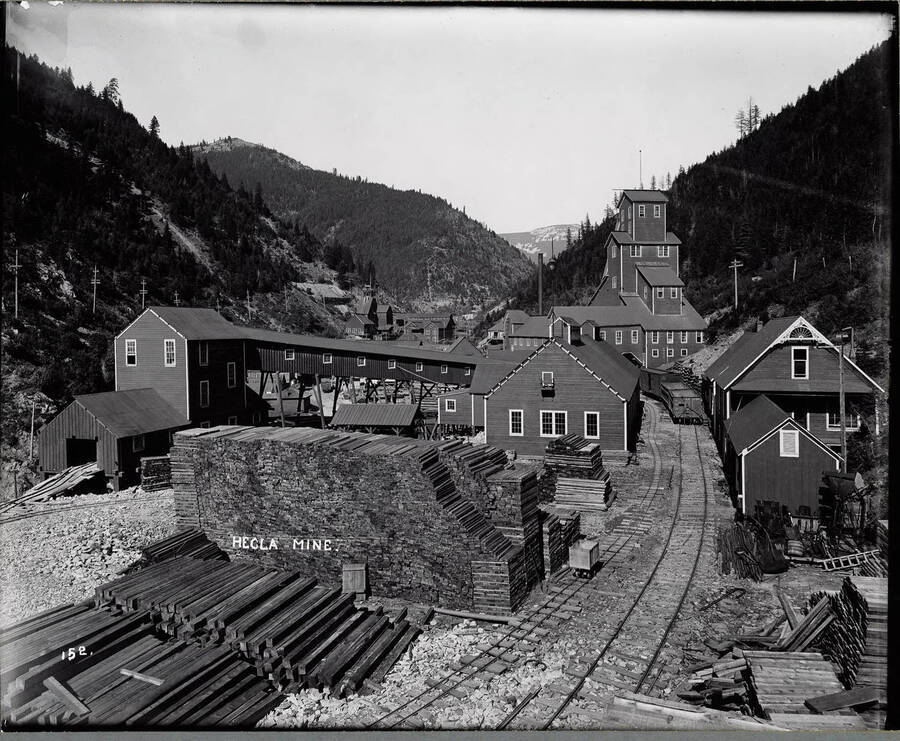 Exterior view of the Hecla Mine in Burke, Idaho. This image shows several buildings, piles of mine timbers and railroad tracks leading through some of the buildings; Caption on front: "Hecla Mine."