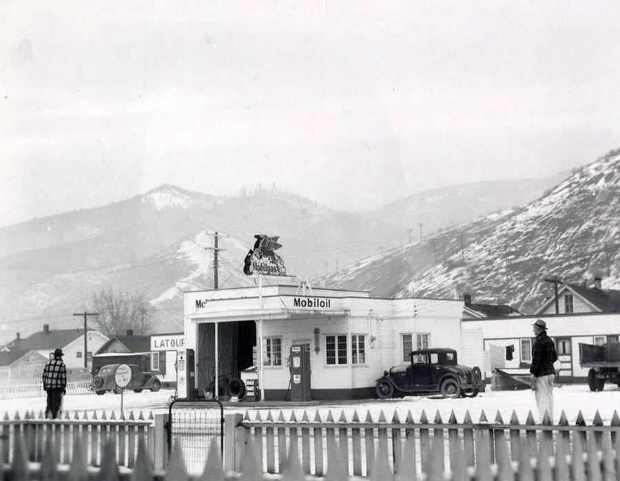 Exterior view of the Mobiloil gasoline station in Wallace, Idaho. There are people standing on the street in front of the station and hills behind it. Taken for Keane & McCann, lawyers.