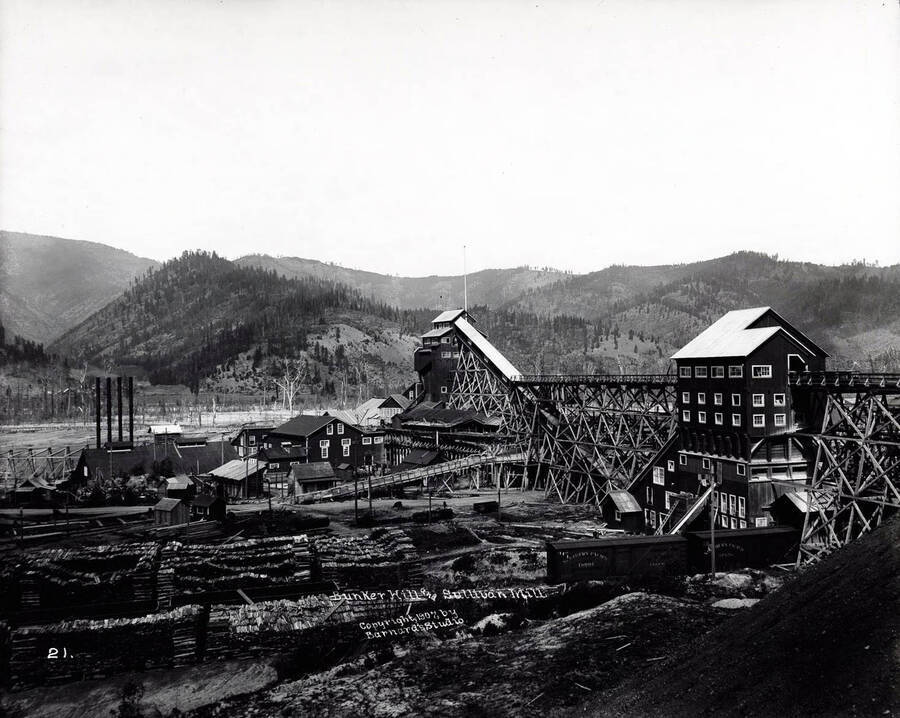 Image is of the Bunker Hill and Sullivan Mill in Kellogg, Idaho [1907]; Piles of lumber, a rail car and various milling buildings are pictured.