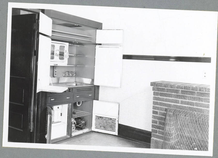 A brick fireplace and hideable kitchenette with its doors open, showing a toaster oven, sink area, a small refrigerator, and shelving, in an upstairs apartment in the Barnard Building in Wallace, Idaho.