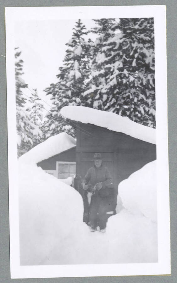 A man standing with a dog outside of a cabin in the snow. Photograph taken for T. Lewis