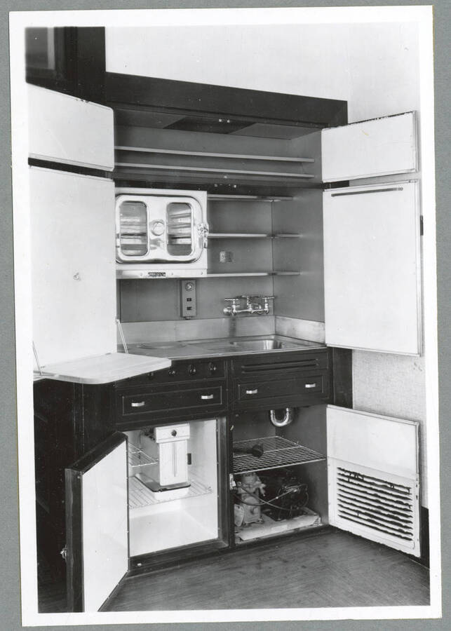 A hideable kitchenette with its doors open, showing a toaster oven, sink area, a small refrigerator, and shelving, in an upstairs apartment in the Barnard Building in Wallace, Idaho.