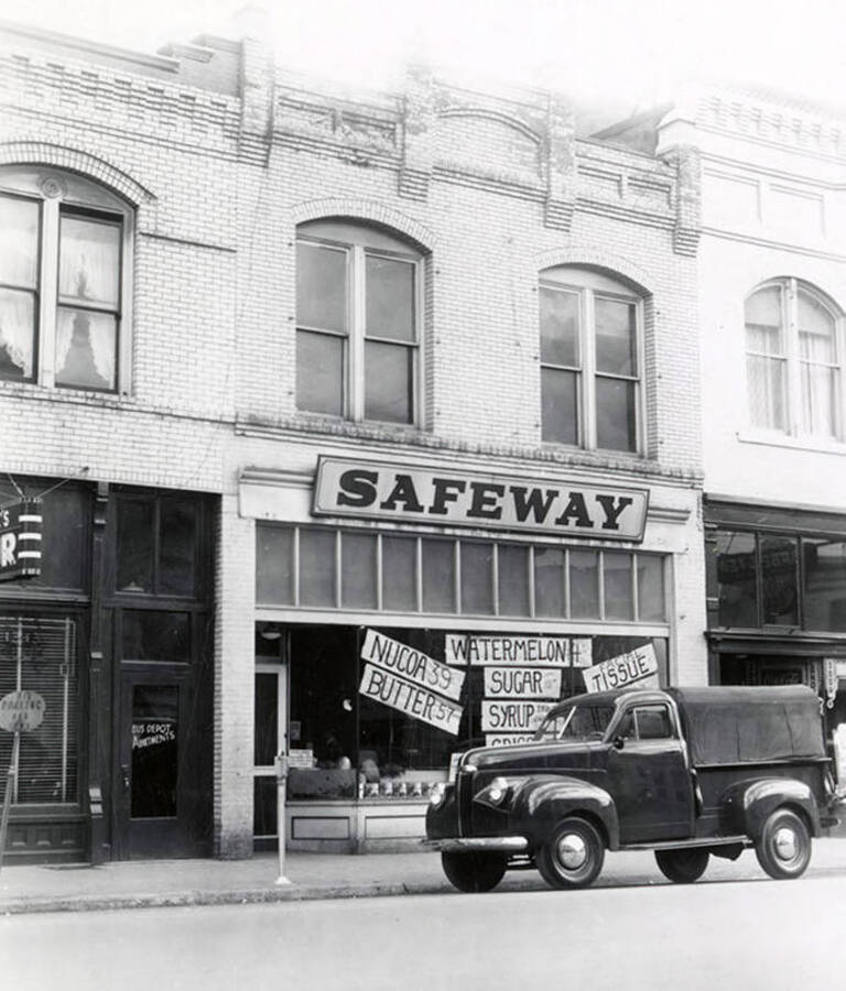 A view from the road of the Safeway store in Wallace, Idaho. Hanging in the window are signs that read, "NUCOA 39," "BUTTER 57," "WATERMELON," "SUGAR," "SYRUP," "CRISCO," "FACIAL TISSUE," "MILK."