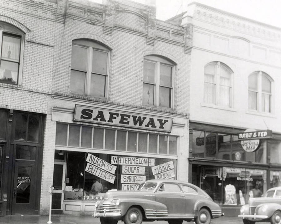 A view from the road of the Safeway store and Babe and Ted's bar in Wallace, Idaho. Hanging in the window of the Safeway are signs that read, "NUCOA 39," "BUTTER 57," "WATERMELON," "SUGAR," "SYRUP," "CRISCO," "FACIAL TISSUE," "MILK."