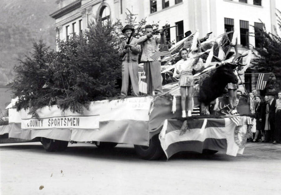 People holding rifles on a float covered in trees and a banner reading "County Sportsmen." On the front, a large moose head is mounted.