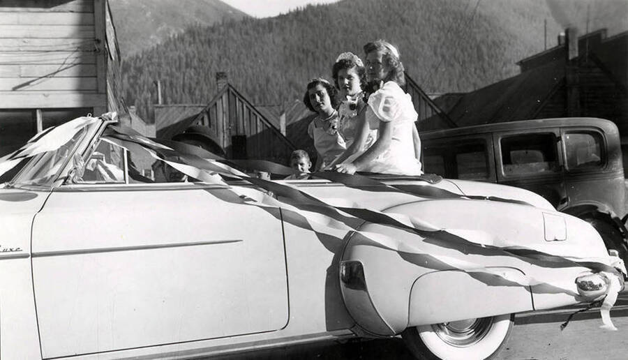 Three women in formal attire sitting atop the backseat of a convertible decorated with streamers during the 49'er Parade in Mullan, Idaho.