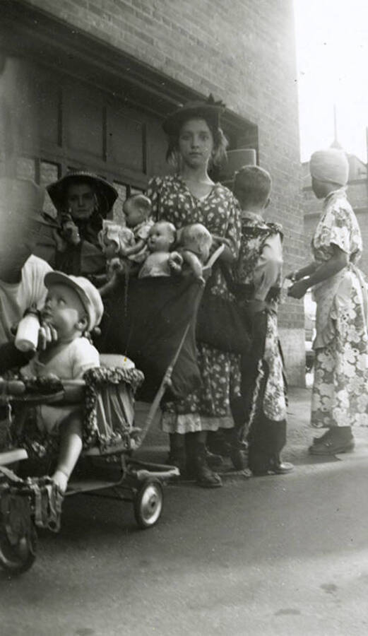 A group of children in costume for the Elks Parade in Wallace, Idaho. The girl in the center is pushing a baby in a stroller with baby dolls.