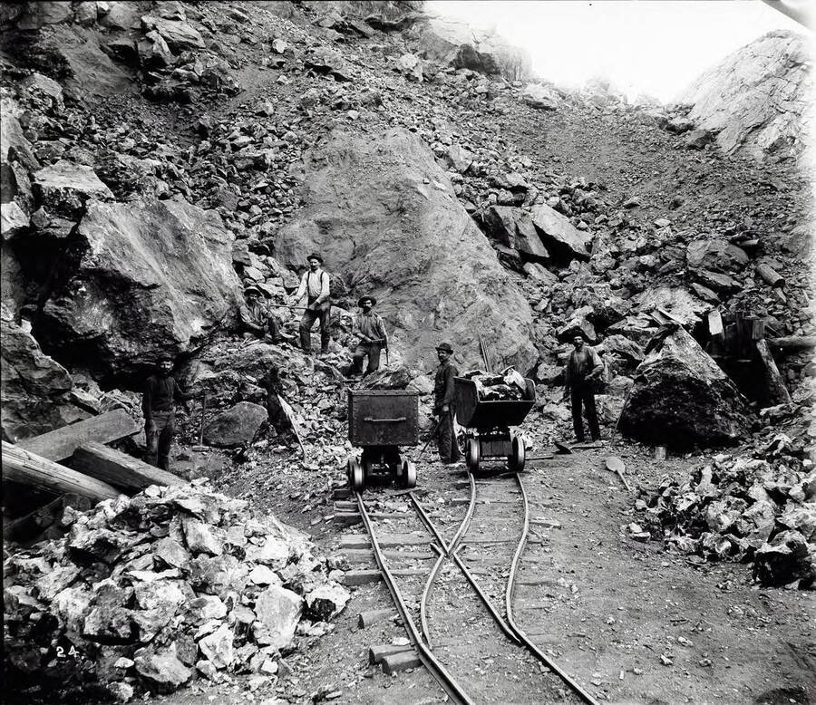 Image shows miners and ore cars at the Bunker Hill "Glory Hole." Caption on front: "Miners work on the original Bunker Hill outcrop." On back of image: "Forty-six years of mining history in the Coeur d'Alene district of northern Idaho came to a close last Thursday, when the Bunker Hill 'Glory Hole' [1886: 1904] Portion of original open 'glory hole.' Location papers were nailed to a big cedar tree on September 10, 1885. Hill and Sullivan Mining and Smelting Company celebrated discovery day at Kellogg. Location papers for the Bunker Hill outcrop, which has been developed into the world's premier lead-silver ore mines, were nailed to a big cedar tree on September 10, 1855, by Noah Kellogg and Phil O'Rourke, veteran prospectors. The picture shows a portion of the original open 'glory hole,' was taken in 1897 [?]. The original location notice was posted near this spot.  The original ore body has been followed downward more than a mile. The deepest workings are said to have exposed ore reserves of great dimension. During the last year an immense, hitherto unknown ore body was discovered on the No. 6 level, almost on the opposite side of the hill from the original discovery. (Spokane Chronicle, September 19, 1931. In Harry Marsh Scrapbook no. 3 page 96, Archives Group 23.)   (In North Idaho Press, Jubilee Edition, June 10, 1958) This picture appears and the caption "miners work on the original Bunker Hill outcrop, 1886)