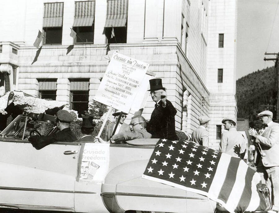 A convertible draped with the American flag carrying men, one with a stovepipe hat, during the Elks Parade in Wallace, Idaho. A sign taped to the side of the car reads "Crusade for Freedom," and a man in the car is holding a sign reading "Crusade for Freedom [...] Vote for Us 1952," and the names Truman Levering, Eisenhower Nelson, Taft Hull, Atchison Eddins, and Abe Gofe.