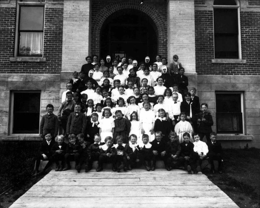 A group of approximately eighty children and nuns posed on the front steps of the Academy of Our Lady of Lourdes school.