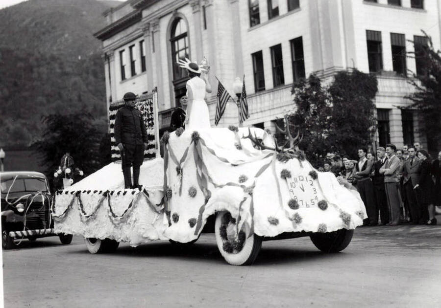 People riding on a float covered in streamers and an American flag. On the front, a deer head is mounted, along with a fake clock.
