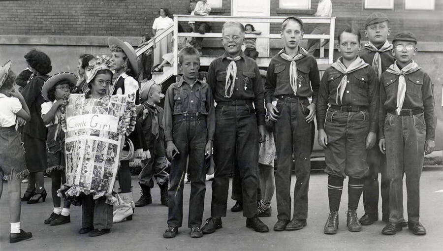 A group of six Boy Scouts posing next to children in costumes during the Mullan 49'er Parade. To the left, a young girl is wearing a costume made out of various comic strips, with "Comic Girl" written across the front.