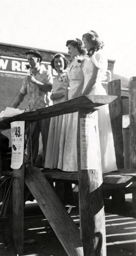 An MC and three women in formal attire standing on a platform during the 49'er Parade in Mullan, Idaho.