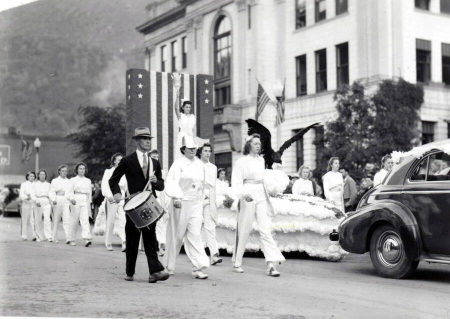 Women in white and a man with a drum marching beside a float during the Elks Roundup Parade in Wallace, Idaho. On the float, a woman dressed as Lady Liberty is holding a torch, and a large black bird is perched on the hood.