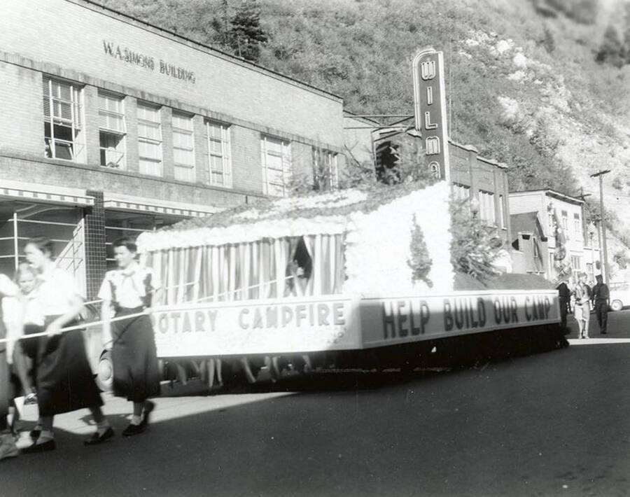 A group of girls pulling a large float past the Wilma Theater during the Elks Parade in Wallace, Idaho. Around the sides of the float is written: "Rotary Campfire / Help Build Our Camp."