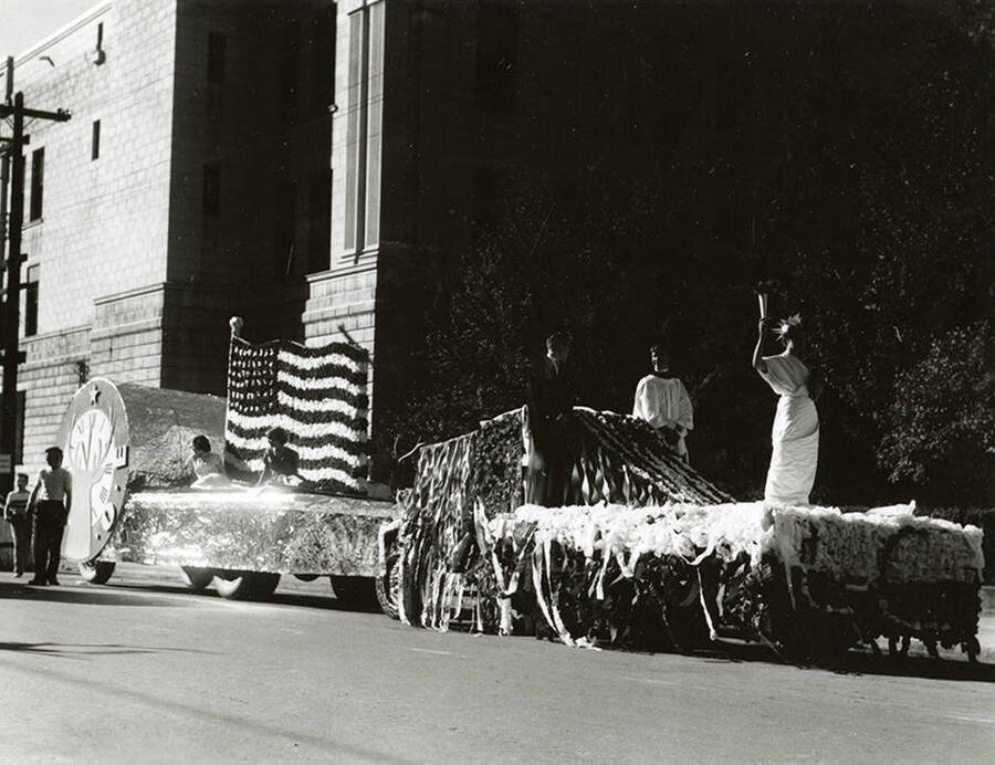 Two men and a woman dressed as Lady Liberty standing atop a float decorated in streamers and a large American flag during the Elks Parade in Wallace, Idaho.