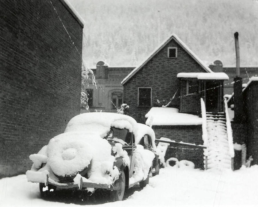 View of a car parked on Cedar Street in Wallace, Idaho covered in snow. Buildings can be seen in the background.