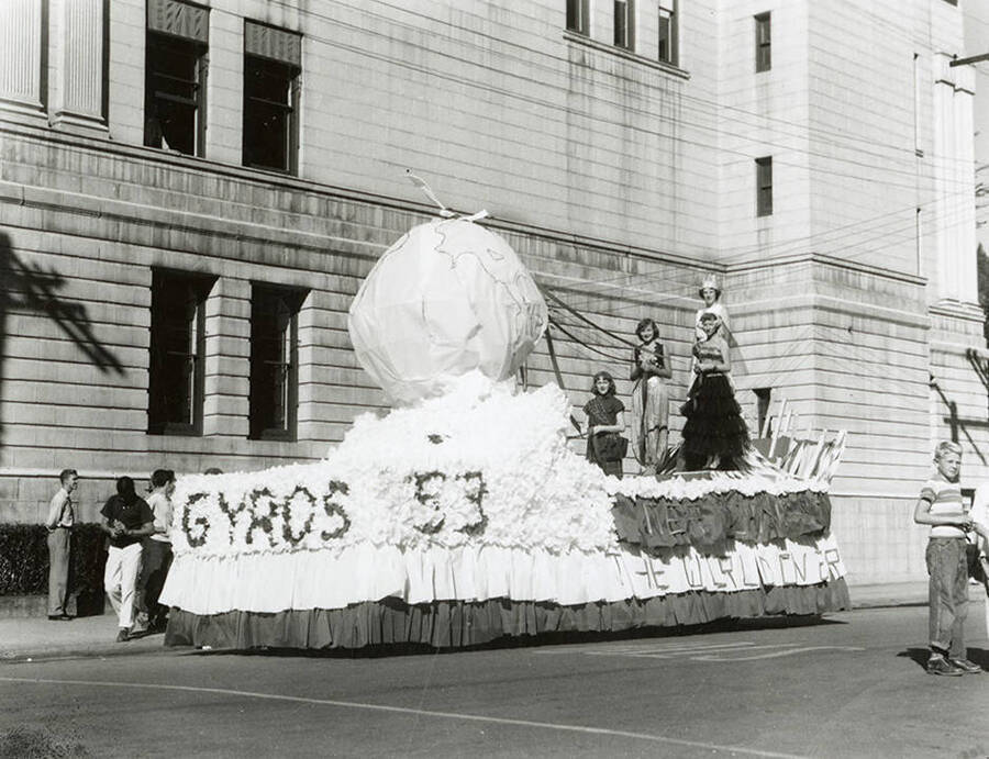 A group of girls standing atop a float decorated with a large, paper globe and signs that read "Gyros 53" and "[unintelligible] The World Over" during the Elks Parade in Wallace, Idaho.