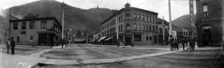 View of street corner in town with people standing on the sidewalks. Panoramic photograph of Wallace, Idaho.