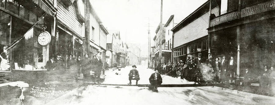 People fishing in Main Street in Burke, Idaho during the Burke flood. Picture comes from a newspaper article. The article reads, "Burke is unique because of the famed street where the river, railroad, and highway are all in the same place. Because of this unusual situation early Canyon Creek floods resulted in an overflow into the street. This March 27, 1913 picture shows adventurous Burke citizens fishing a board stretched across the creek while the rampaging Canyon Creek rushes through the town."