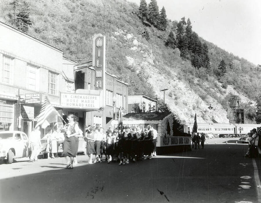 A group of girls pulling a large float past the Wilma Theater during the Elks Parade in Wallace, Idaho. Around the sides of the float is written: "Rotary Campfire / Help Build Our Camp."