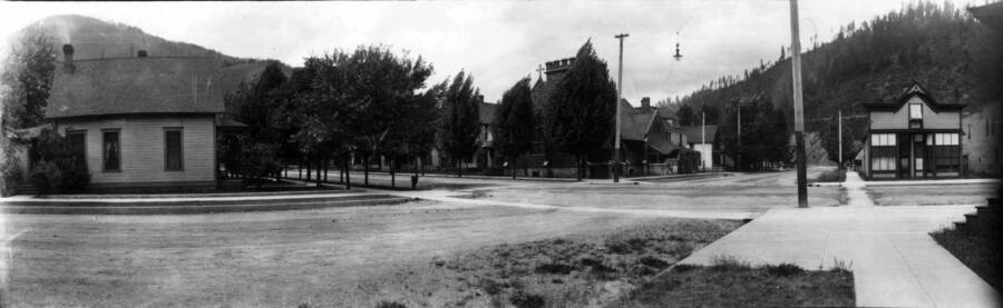 View of a street corner with houses and the Holy Trinity Episcopal Church behind some trees. Panoramic photograph of Wallace, Idaho.