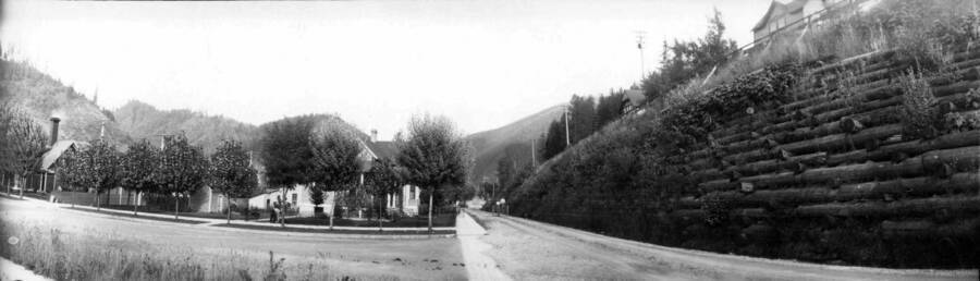 View of tree lined dirt roads and houses. Two boys are riding a bike down the road. Panoramic photograph of Wallace, Idaho.