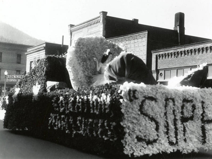 A woman sitting atop a large float during the Elks Parade in Wallace, Idaho. Flowers are arranged around the sides of the float to read: "Education [unintelligble] Success / SOPH [unintelligble]"