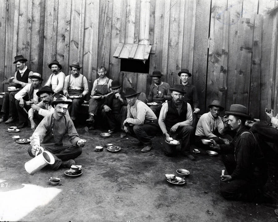 Image shows a group of miners eating at the Bull Pen in Kellogg, Idaho [1899]; Tin cups and plates were used. One man is pouring his drink from a tin pitcher.