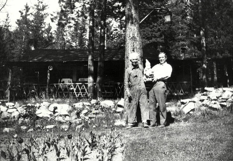 Two men holding a gnome and standing against a rock fence at Tobs cabin near Wallace, Idaho.