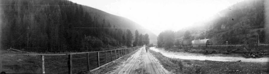 A man walking down a dirt road with a farm and stream nearby. Panoramic photograph of Wallace, Idaho.
