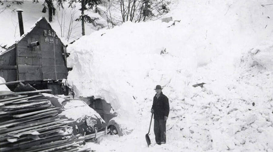 A man standing with a shovel beside his snow-covered house and cars in historical Yellow Dog, Idaho, after a snow slide.