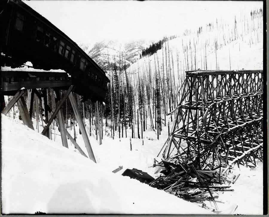 Image shows a passenger car dangling from the broken edge of the "S" bridge trestle. The bridge was destroyed by a heavy snow slide. The shadow of the photographer can be seen in the lower half of the photo.