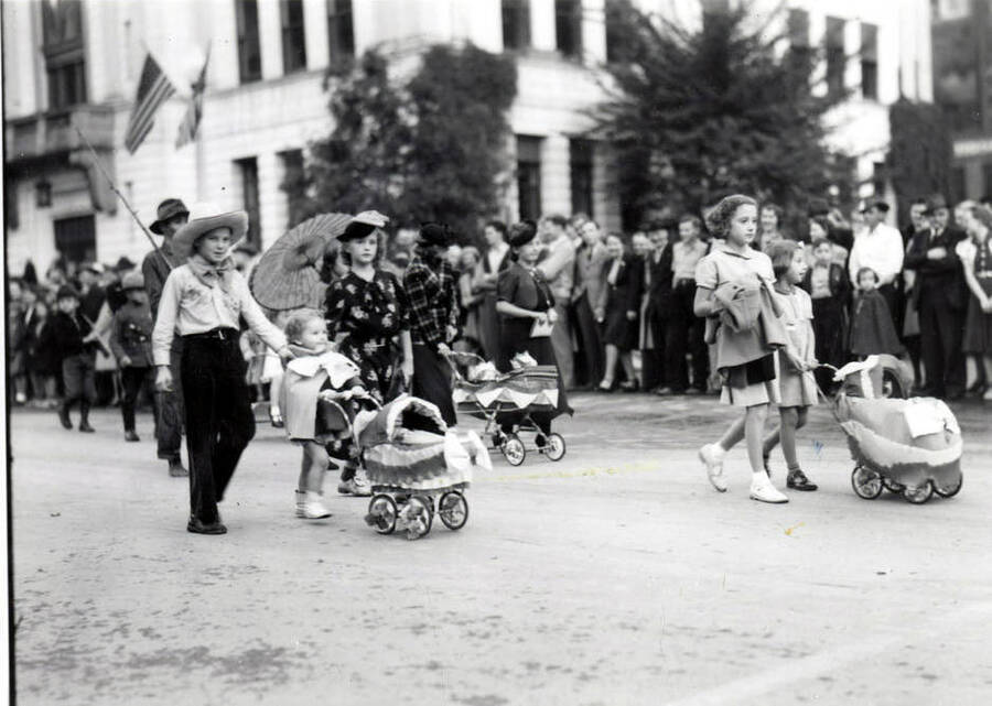 Children in costume walking along with parasols and baby carriages during the Elks Roundup Parade in Wallace, Idaho.
