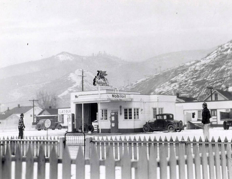 Exterior view of the Mobiloil gasoline station in Wallace, Idaho. There are people standing on the street in front of the station and hills behind it. Taken for Keane & McCann, lawyers.