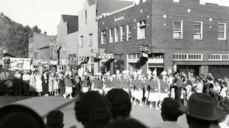 Women wearing shako hats and marching down the street during the 49'er Parade in Mullan, Idaho. To the left, a large sign is mounted on the roof of a car, reading "Win THIS CAR!"
