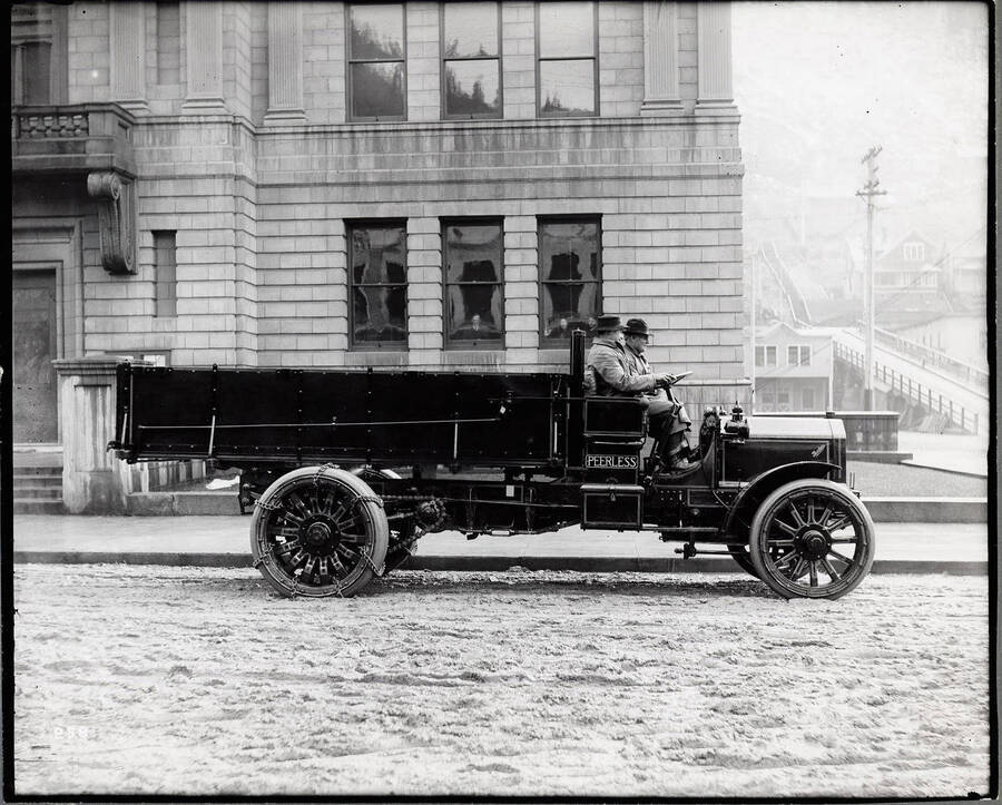 Image of a 1915 [?] Peerless 4-ton truck with two men in the cab, parked in front of the Shoshone County Court House in Wallace. A man and two women can be seen peering out of two of the Court House windows.