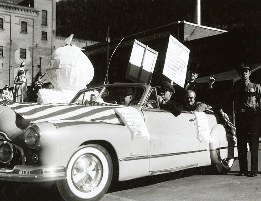 A convertible draped with the American flag carrying men, one with a stovepipe hat, during the Elks Parade in Wallace, Idaho. A sign taped to the side of the car reads "Crusade for Freedom," and a man in the car is holding a sign reading "Crusade for Freedom [...] Vote for Us 1952," and the names Truman Levering, Eisenhower Nelson, Taft Hull, Atchison Eddins, and Abe Gofe.
