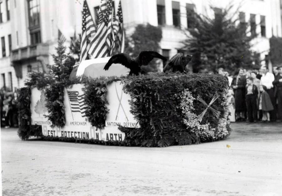 A float covered in tree branches and American flags, with a . Written on the side beneath images of trees, an American flag, and two sabers, is, "Conservation / Americanism / National Defense / Best Protection On Earth." Two large black birds are on top of the float.