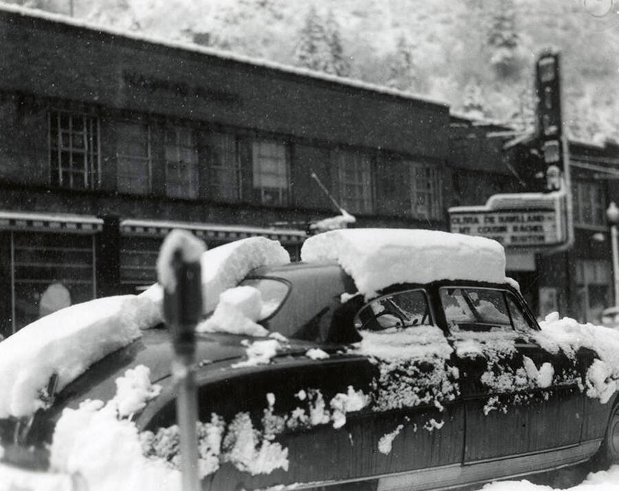 View of a car parked on Cedar Street in Wallace, Idaho covered in snow. Buildings can be seen in the background.