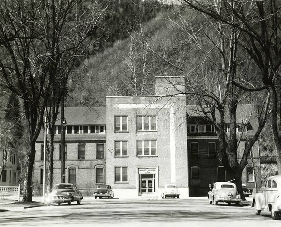 Exterior view of the hospital in Wallace, Idaho. Cars are seen parked out front and a mountain is in the distance.