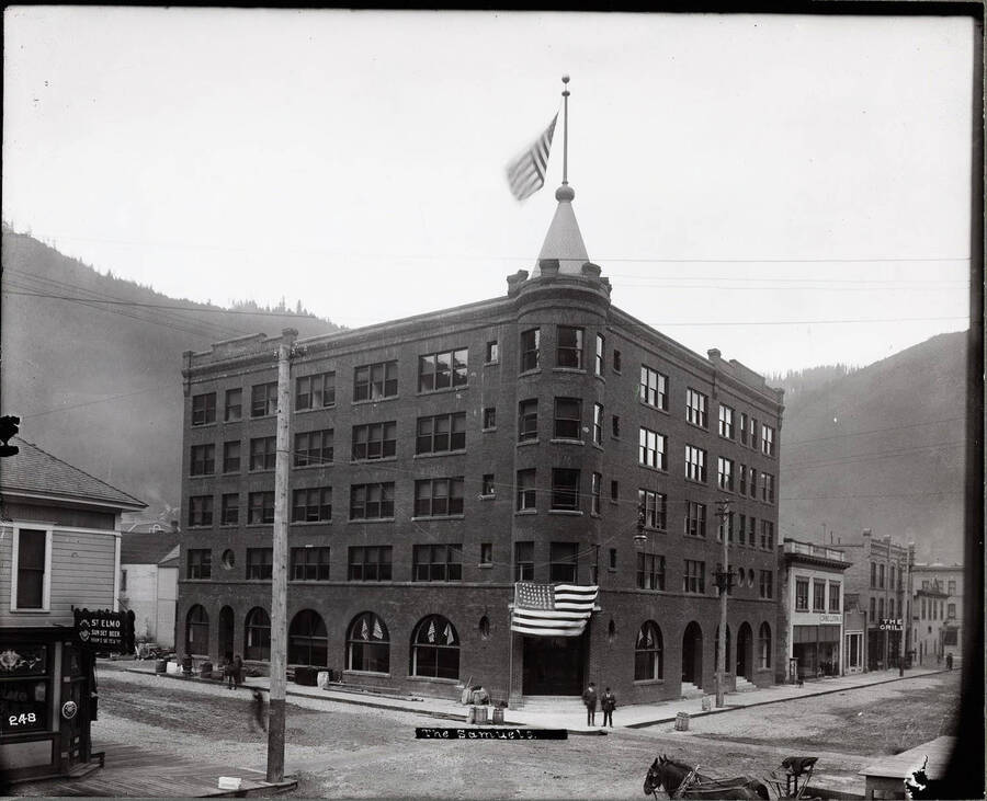 Exterior of the Samuel's Hotel with one American flag in front of the entrance and one flying from its turret. Two men are standing in front of the building.