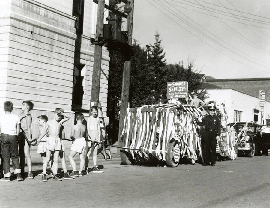 A group of boys pulling a float decorated in streamers in the Elks Parade in Wallace, Idaho. A police officer is standing beside the float.
