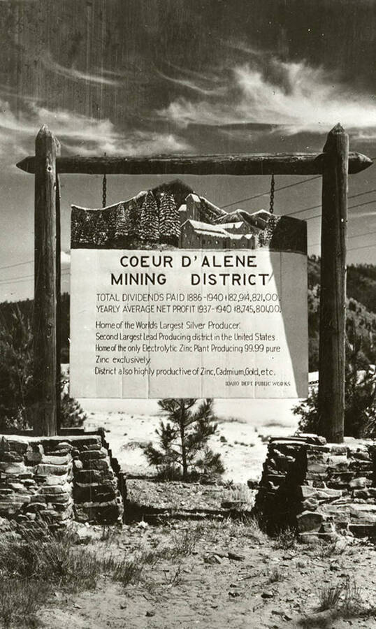 A sign hanging from a log frame reading: "Coeur d'Alene Mining District / Total dividends paid 1886-1940 $182,914,821.00 / Yearly average net profit 1937-1940 $8,745,801.00 / Home of the Worlds Largest Silver Producer. Second Largest Lead Producing district in the United States. Home of the only Electrolytic Zinc Plant Producing 99.99 pure Zinc exclusively. District also highly productive of Zinc, Cadmium, Gold, etc. / Idaho Dept. Public Works"