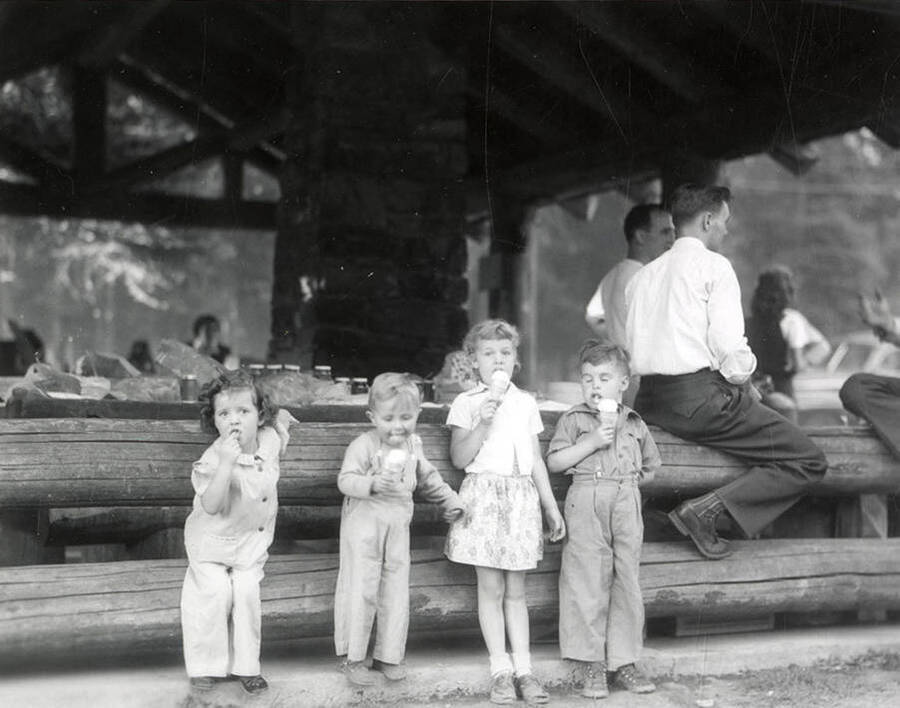 A few children eating ice cream during Wallace Jaycee's picnic in Pottsville, Idaho. Some men sit on a log behind the children.